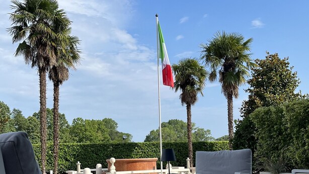 An outdoor seating area with white chairs and a table, a flagpole with a flag, palm trees, and a hedge in the background. - Hotel Poppi (Mira)