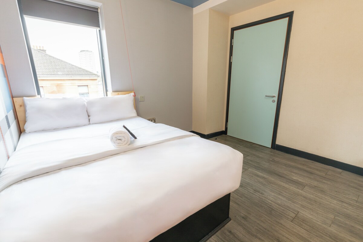 A neatly made bed with white linens and a rolled towel, a wooden floor, and a closed door.