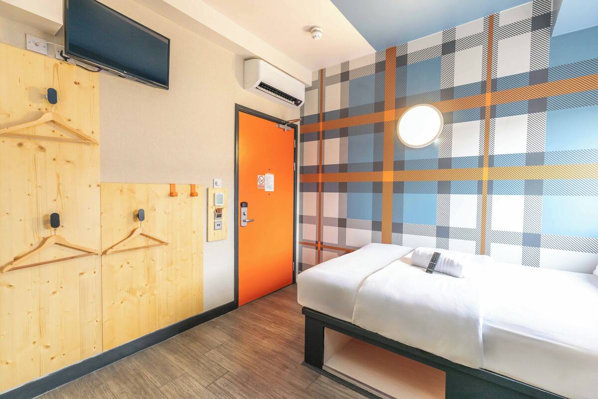Een hotelkamer met een bed, een televisie, een garderobe met houten kleerhangers, een oranje deur en een wand met een geruit patroon.