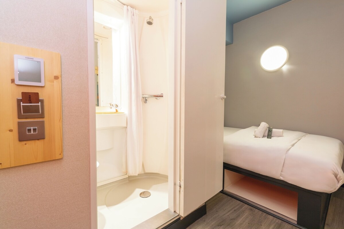 A hotel room with a shower, a bed with a pillow and a remote control, and a wall-mounted light fixture.