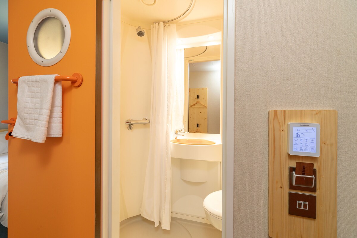 Une salle de bain avec un lavabo blanc, des toilettes et un rideau de douche. On y trouve un porte-serviettes avec des serviettes blanches et un thermostat numérique Samsung fixé au mur.
