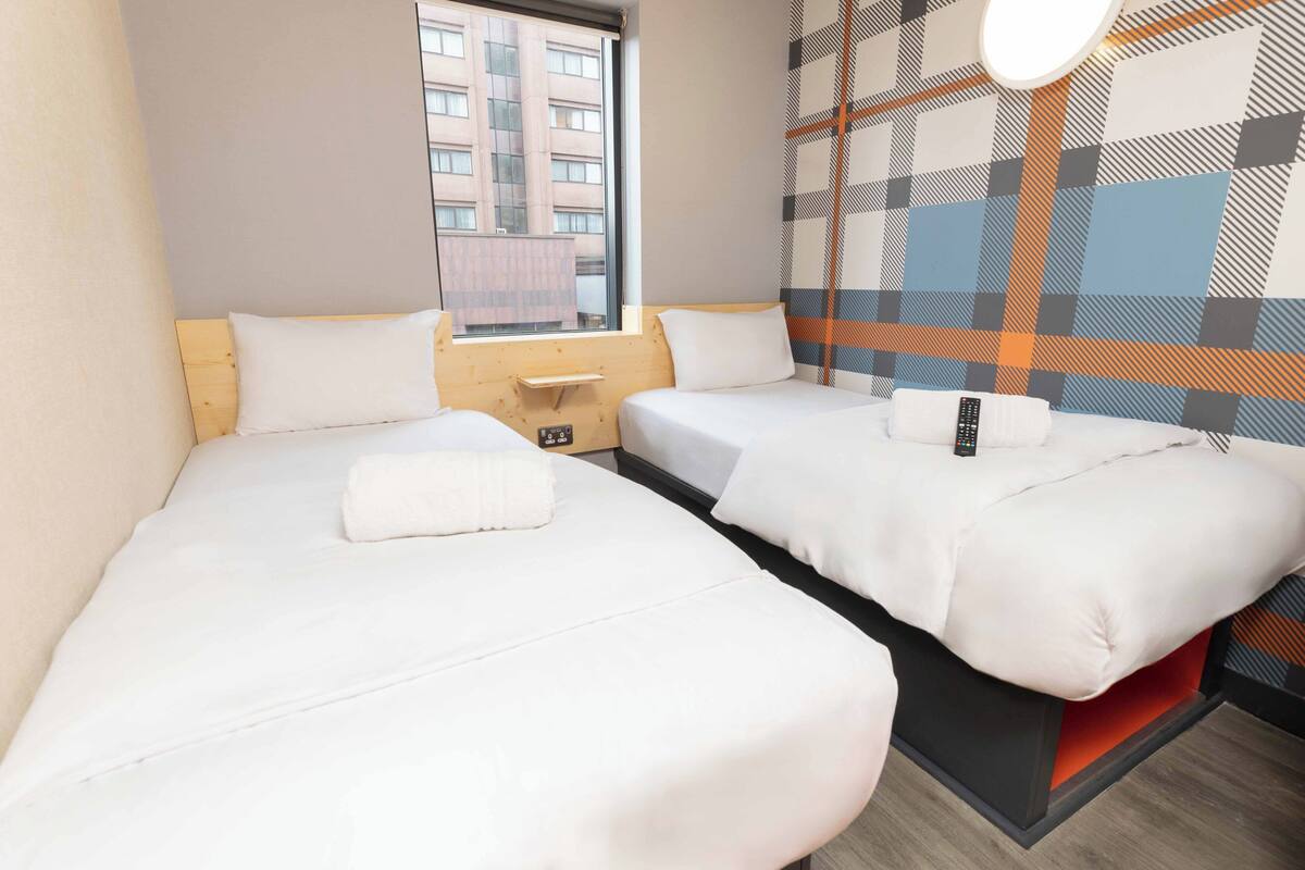 Hotelkamer met twee bedden, een raam, een wandlamp en uitzicht op een gebouw.