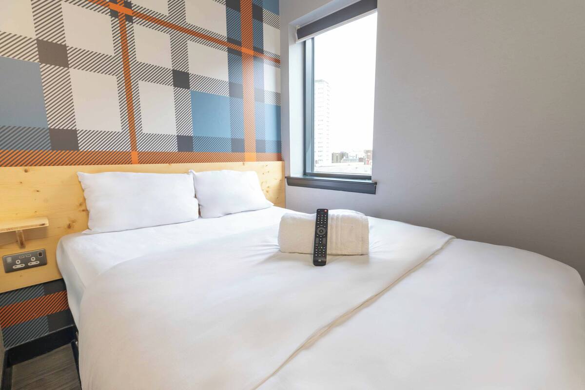 Een hotelkamer met een groot bed, een afstandsbediening en een raam met uitzicht op de stad.