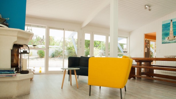 A spacious room with a wooden floor, a black sofa, a yellow chair, a small table, and a painting of a lighthouse.
