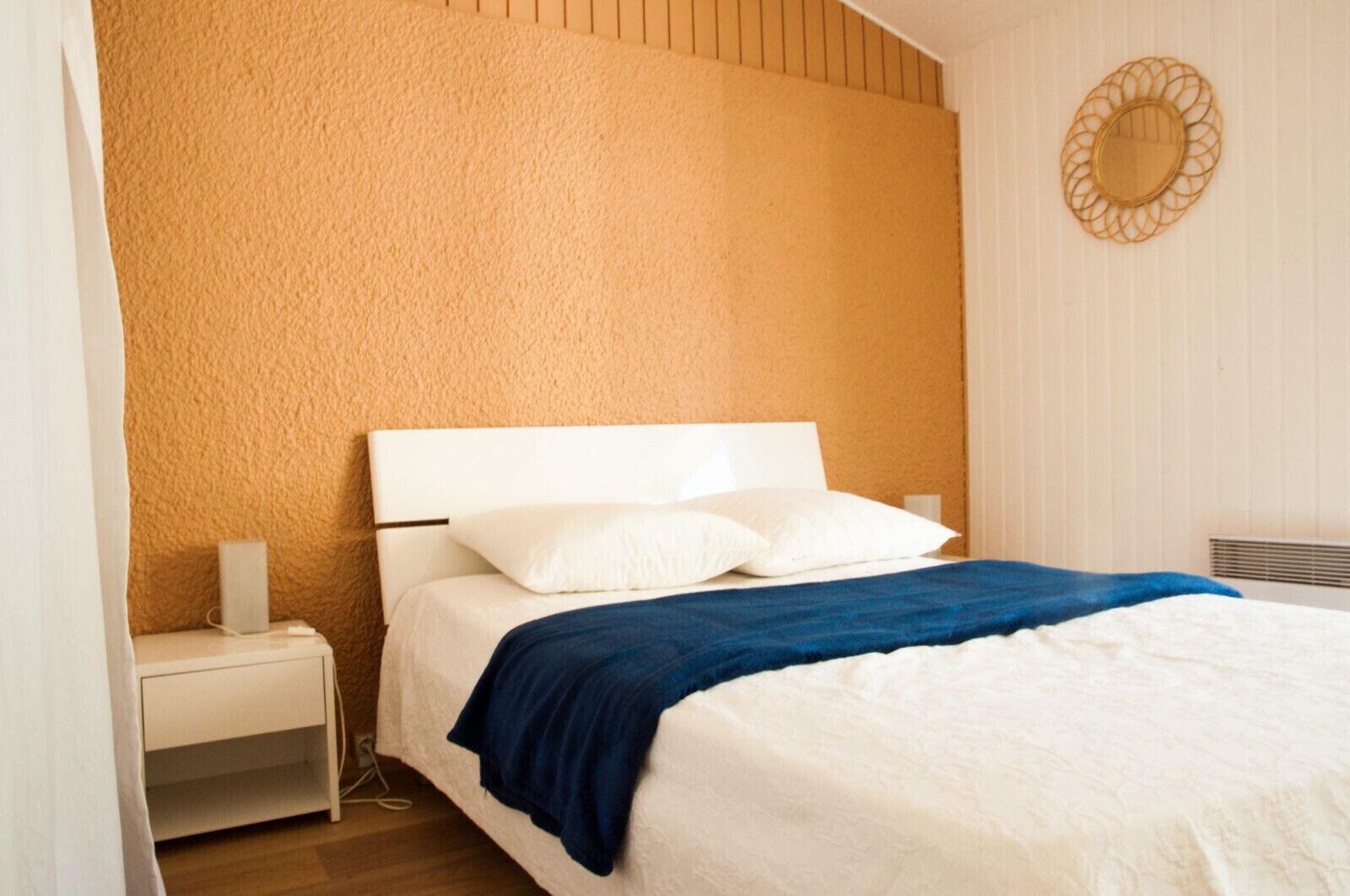 A bedroom with a white bed, blue blanket, and white pillows. There is a white nightstand with a lamp and a power outlet. The wall is textured and painted orange.