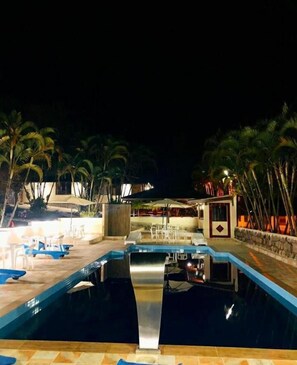 A swimming pool with lounge chairs and umbrellas, surrounded by palm trees and a building with a stone wall. - Hotel Fazendinha (Itatiaia)