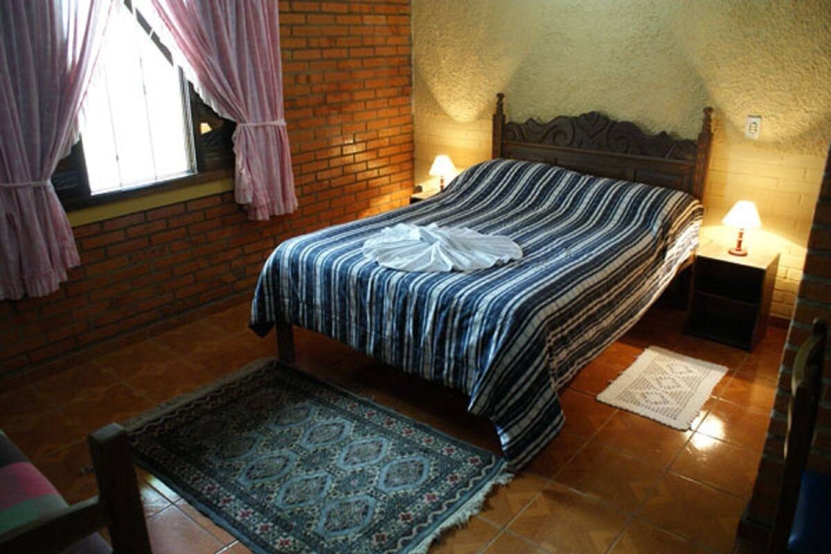 A bedroom with a bed, two bedside lamps, a window with curtains, a patterned rug, and a brick wall.