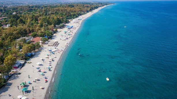 Vista aérea de una playa con aguas azules cristalinas, una extensa franja de arena y una hilera de árboles y edificios a lo largo de la costa.