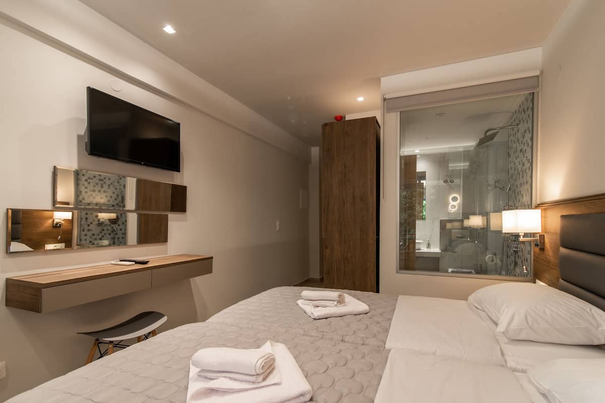 A modern hotel room with a large bed, a desk with a chair, a TV mounted on the wall, and a large window with a view of the bathroom.