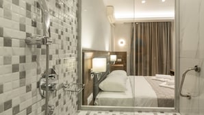 Un baño de hotel moderno con cabina de ducha de vidrio, pared con azulejos y una habitación visible con cama y mesita de noche.