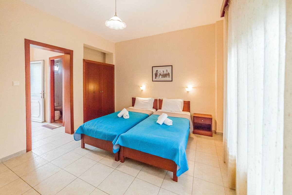 A hotel room with two beds, a wooden wardrobe, a framed picture on the wall, and a door leading to another room.