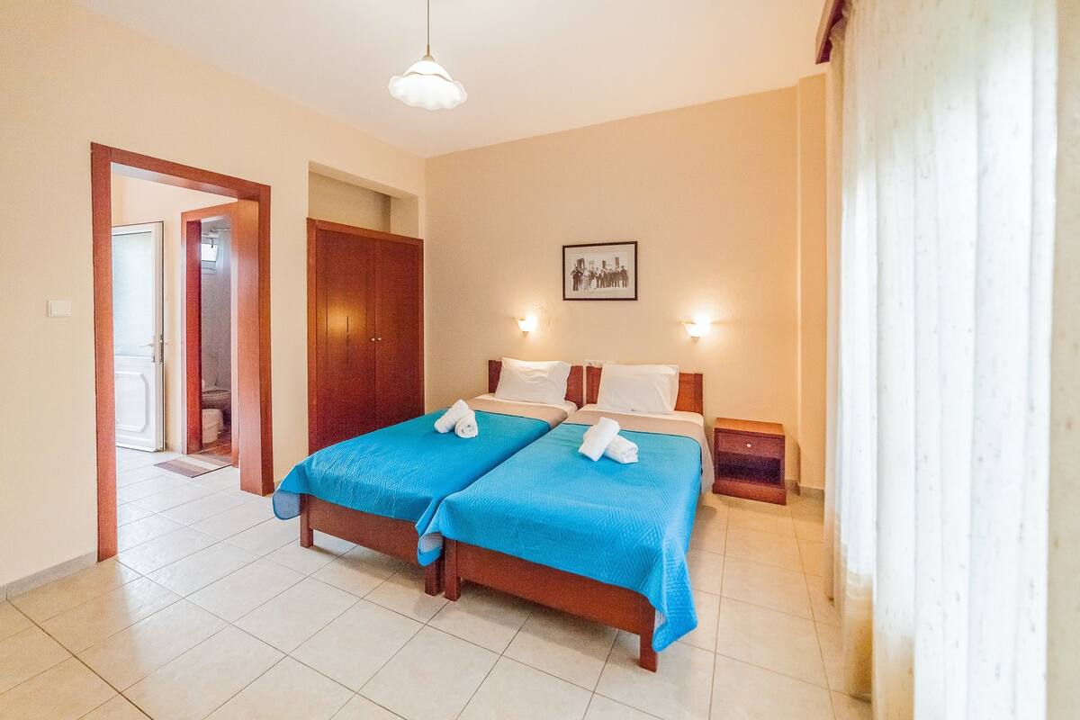 A hotel room with two beds, a wooden wardrobe, a framed picture on the wall, and a door leading to another room.