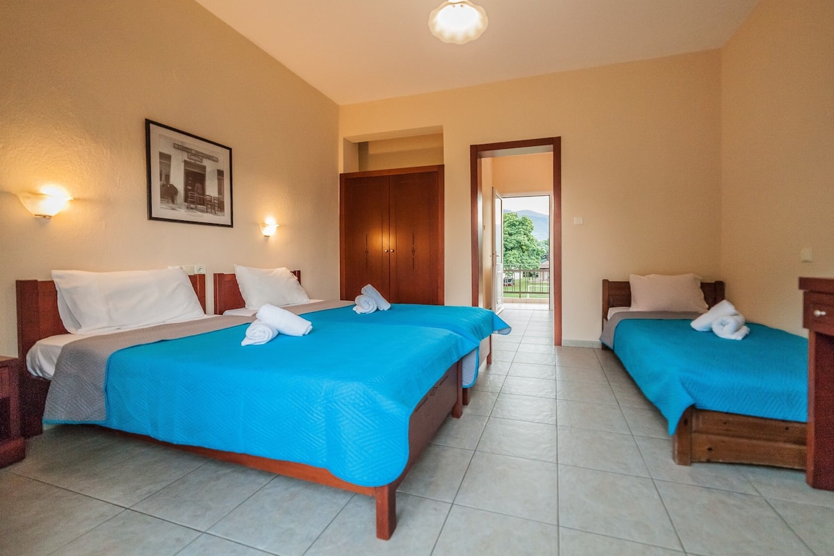 A hotel room with two beds, each with a blue bedspread and white pillows. There is a framed picture on the wall and a door leading to a balcony.