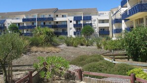 A multi-story hotel with balconies, a garden area with shrubs and a wooden fence, and a clear blue sky. - Appartement Bleu Azur by Agence Bru - 2051 (Lacanau)