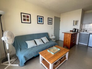 A compact living space with a sofa, a coffee table, a fan, a kitchenette, and framed pictures on the wall.