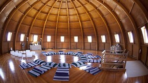 Une vaste pièce circulaire dotée d’un plafond en forme de dôme en bois et d’un puits de lumière central. La pièce est recouverte d’un parquet en bois et meublée de chaises et de tables blanches disposées en demi-cercle.