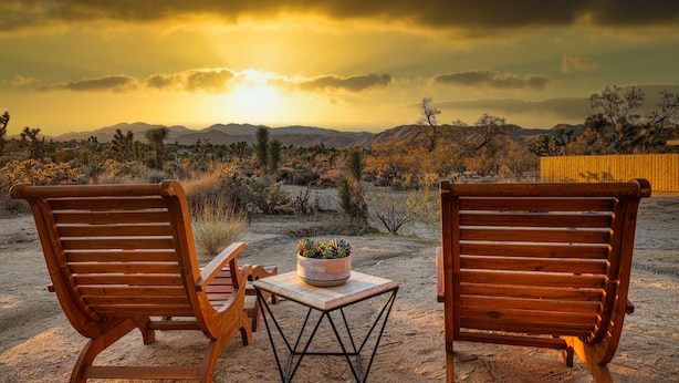 Deux chaises en bois, une petite table et un pot de fleurs placés entre elles, le tout installé sur un sol sablonneux, avec un paysage désertique et des montagnes en arrière-plan.