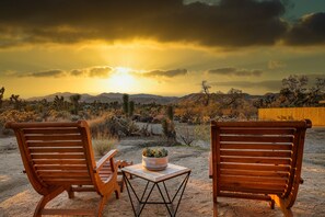 Two wooden chairs with a small table and a potted plant between them, set on a sandy ground with a desert landscape and mountains in the background. - Panoramic Views & Hot Tub! Joshua Tree Sunset Vista (Yucca Valley)