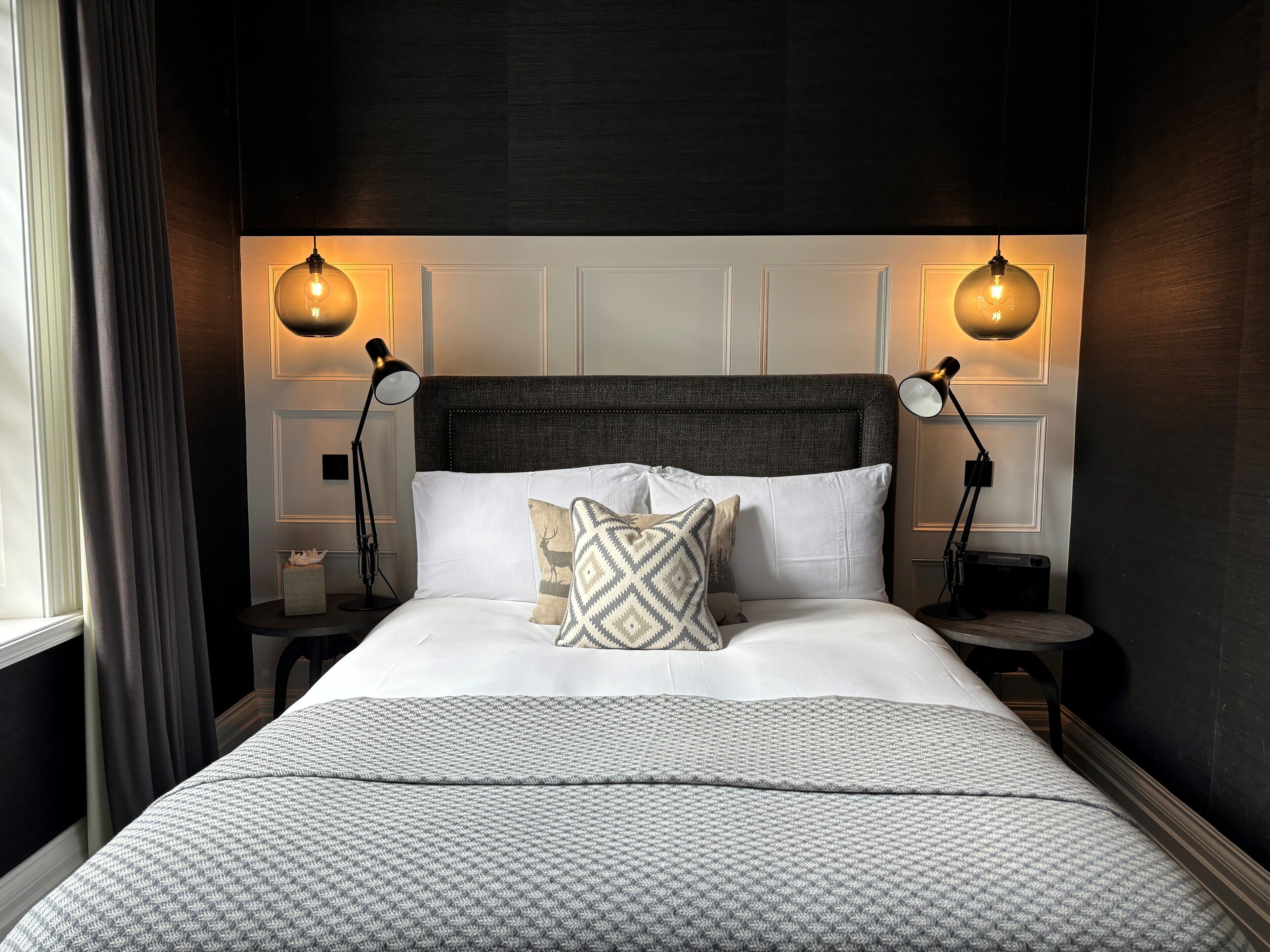Un lit bien fait, avec du linge de lit blanc et des coussins à motifs, flanqué de deux lampes de chevet.