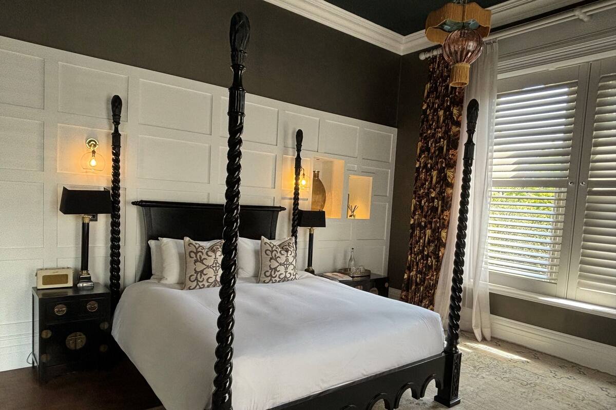 A four-poster bed with a dark wooden frame, white bedding, and decorative pillows. There are two bedside tables with lamps and a small vase on one. The room has a patterned carpet and a window with blinds.