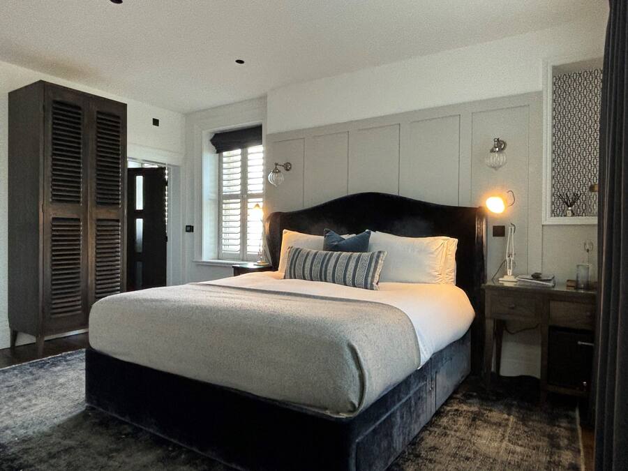 Ein großes Bett mit dunklem Kopfteil, ein Nachttisch mit Lampe und ein Fenster mit Rollläden.