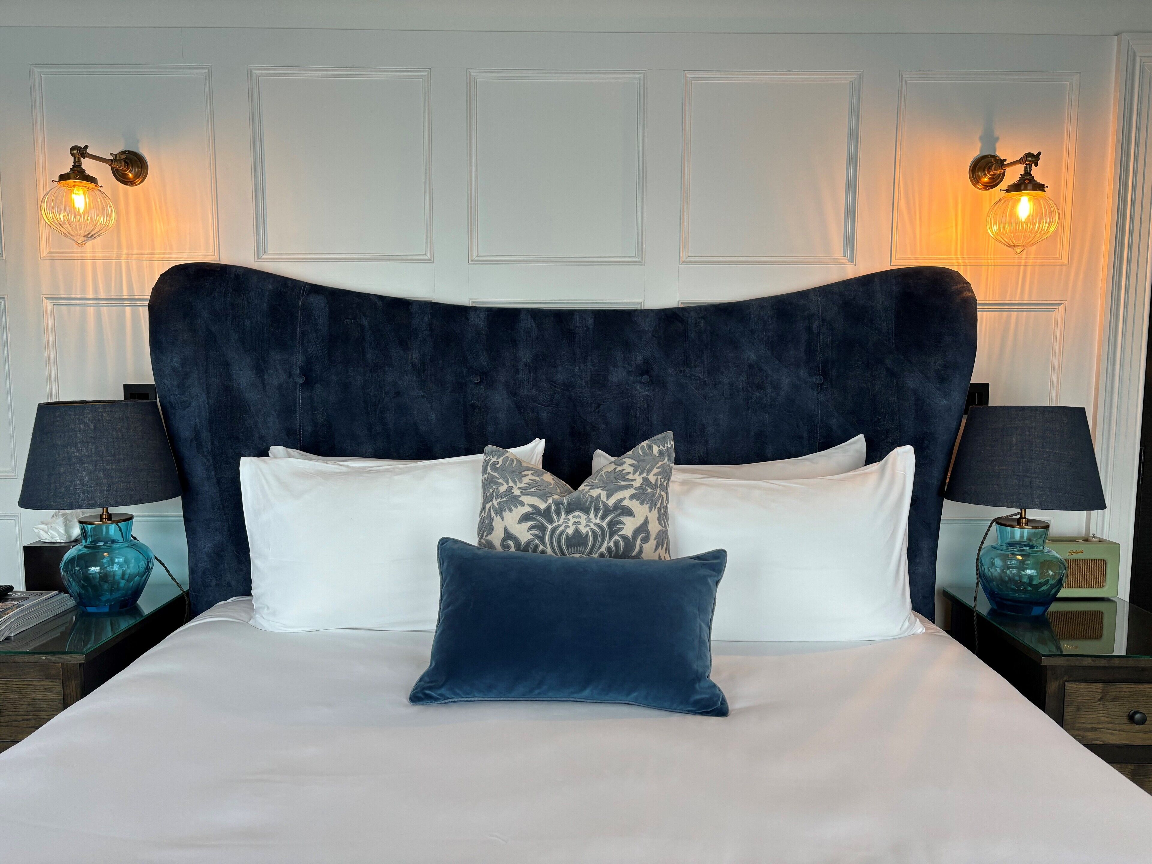 A bed with a dark headboard, white bedding, and decorative pillows. There are two bedside tables with lamps and a blue glass vase on each.