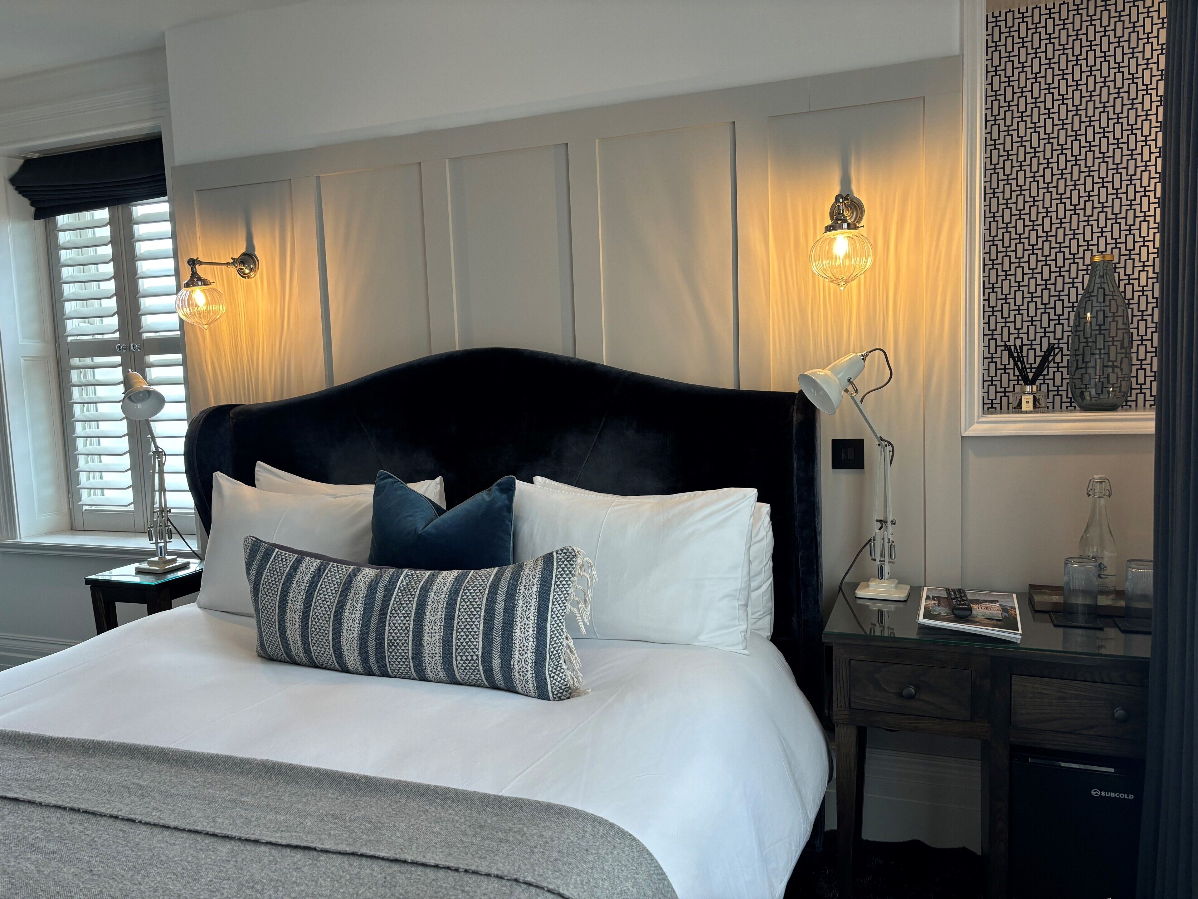 Una cama bien hecha con almohadas blancas y estampadas, un cabecero oscuro y lámparas de noche.