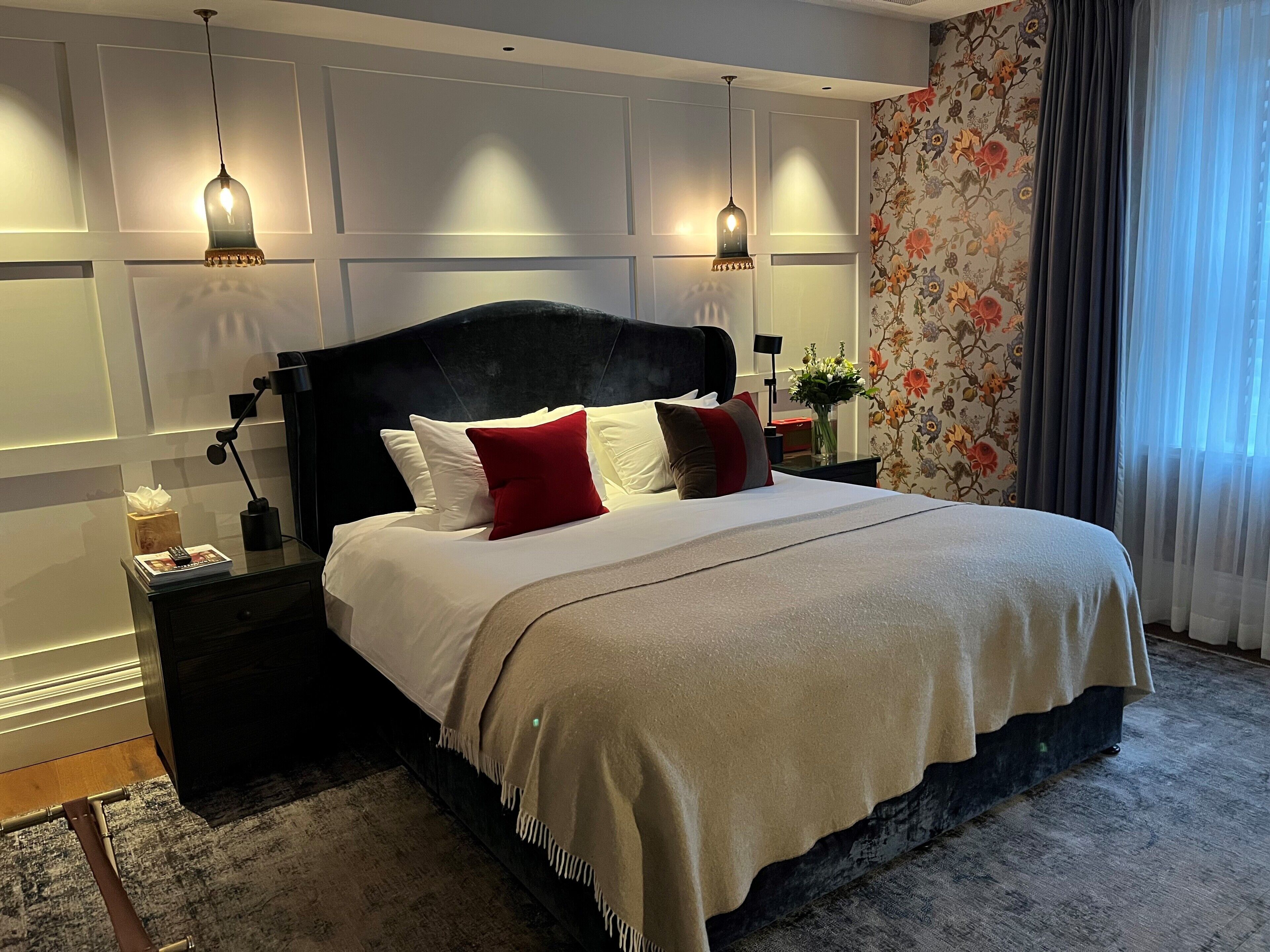 A modern hotel room with a large bed, two bedside lamps, a nightstand, and a floral wallpaper.