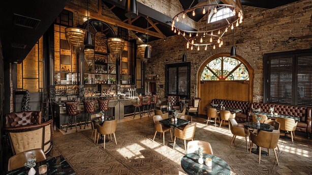 A bar area with wooden beams, a brick wall, and a large arched window. Tables and chairs are arranged for dining. - Manor House Lindley (Huddersfield)