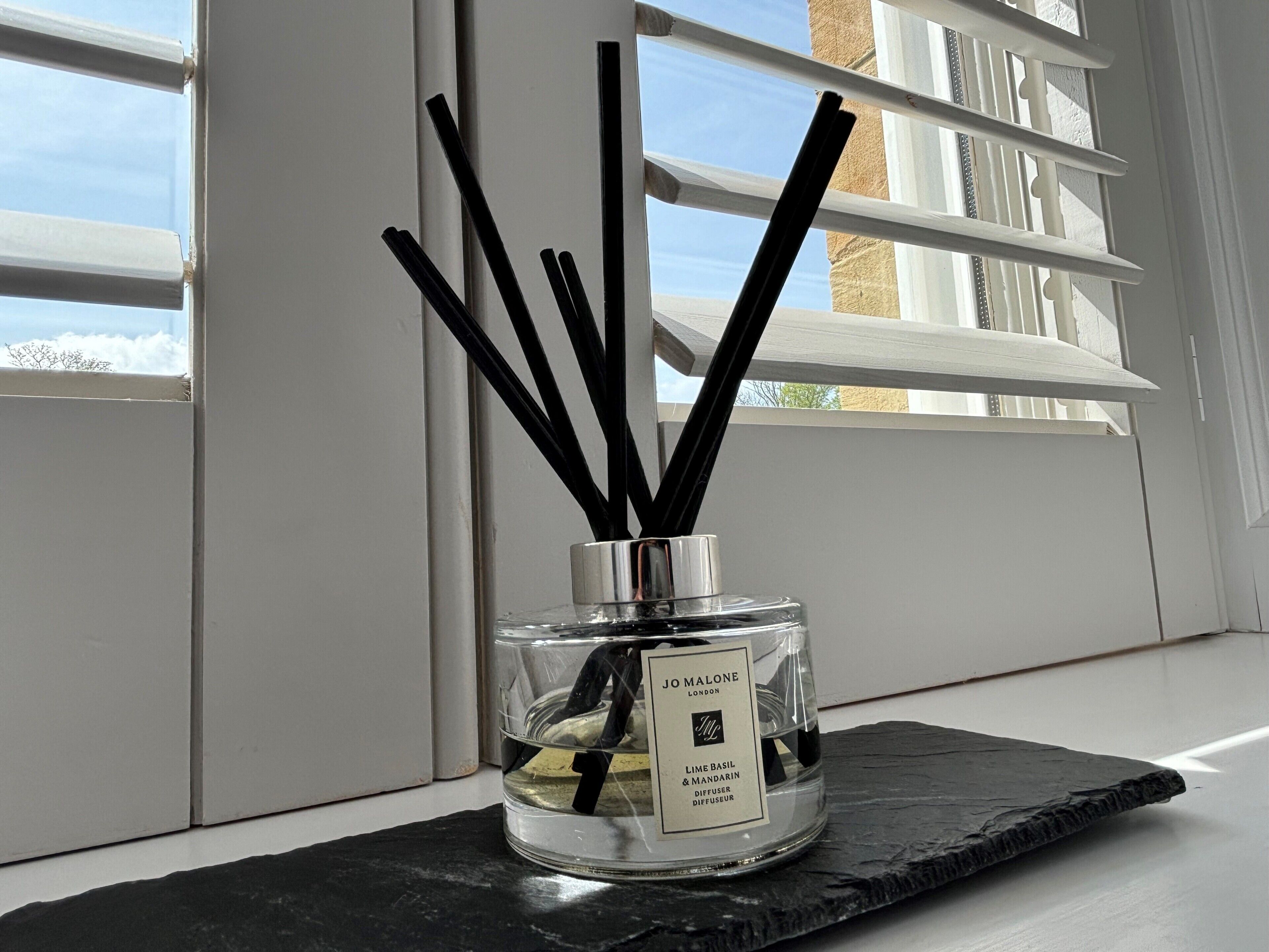 Un difusor de vidrio con cañas negras, etiquetado 'JO MALONE LONDON LIME BASIL & MANDARIN' sobre una superficie de pizarra negra.