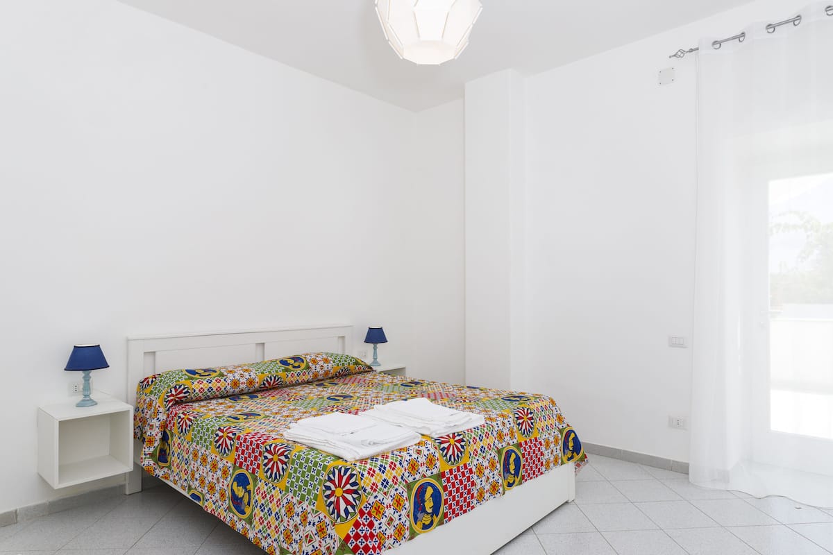 Une chambre à coucher avec une couvre-lit aux couleurs vives, des murs blancs et une fenêtre avec un rideau.