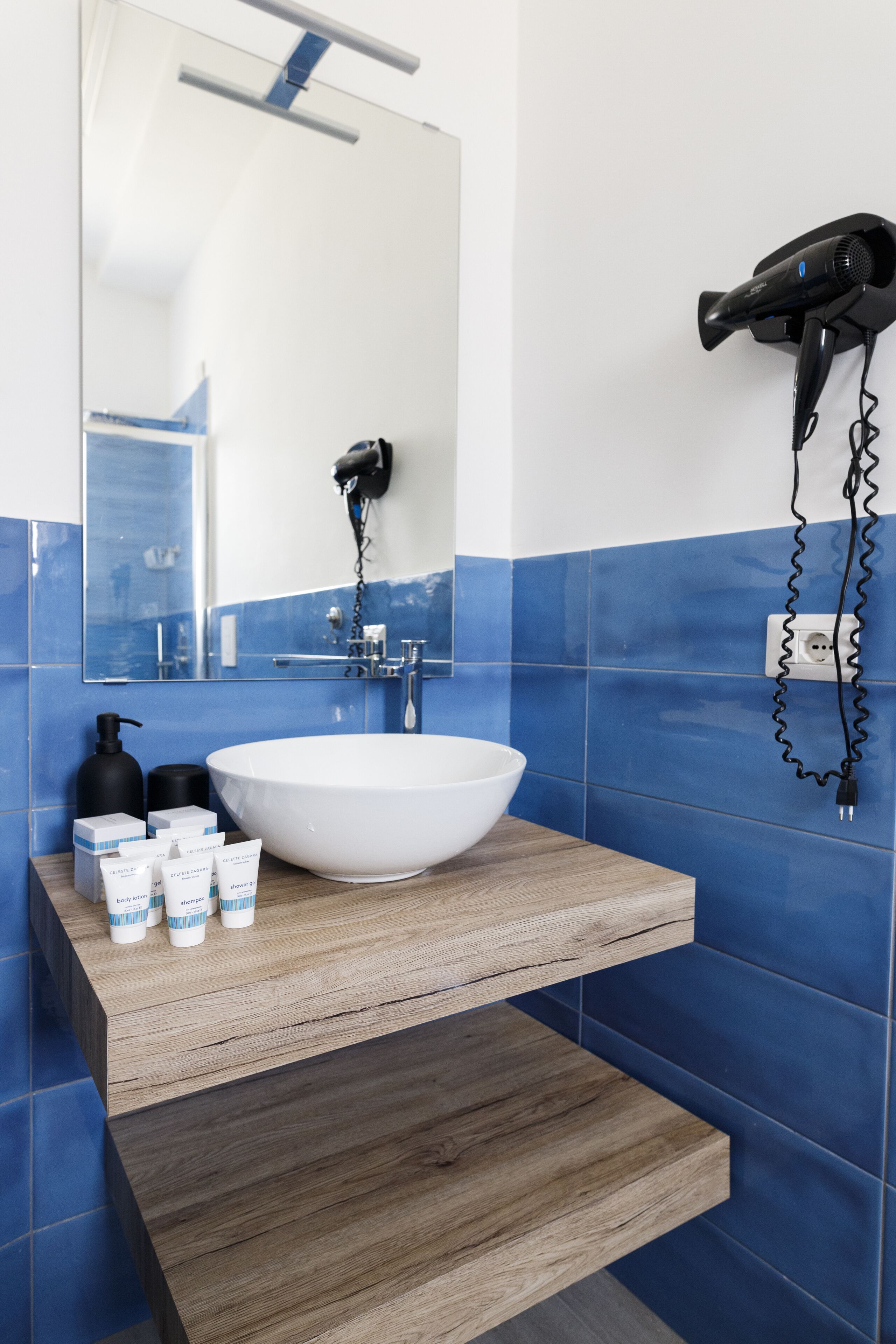 A bathroom with a white sink, blue tiles, a wooden vanity, and a hairdryer mounted on the wall.