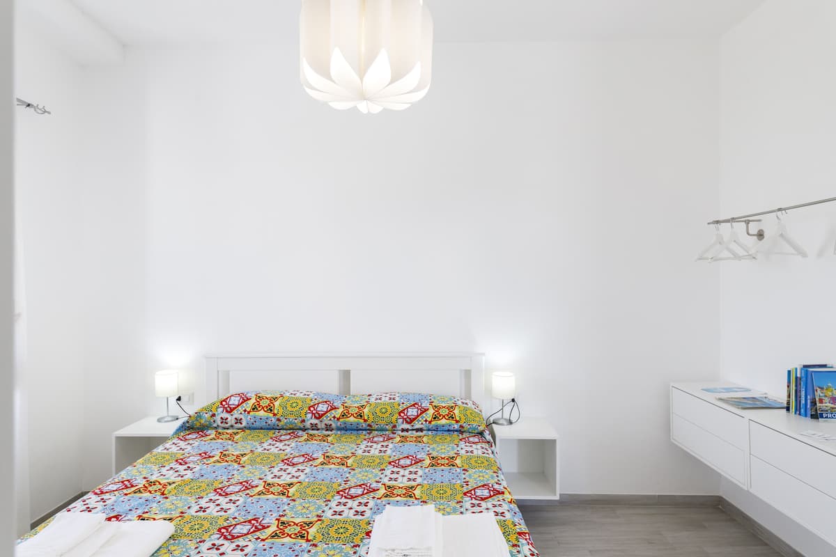 Une chambre à coucher avec une couvre-lit aux couleurs vives, des lampes de chevet, une table de chevet blanche et un petit bureau avec des livres.