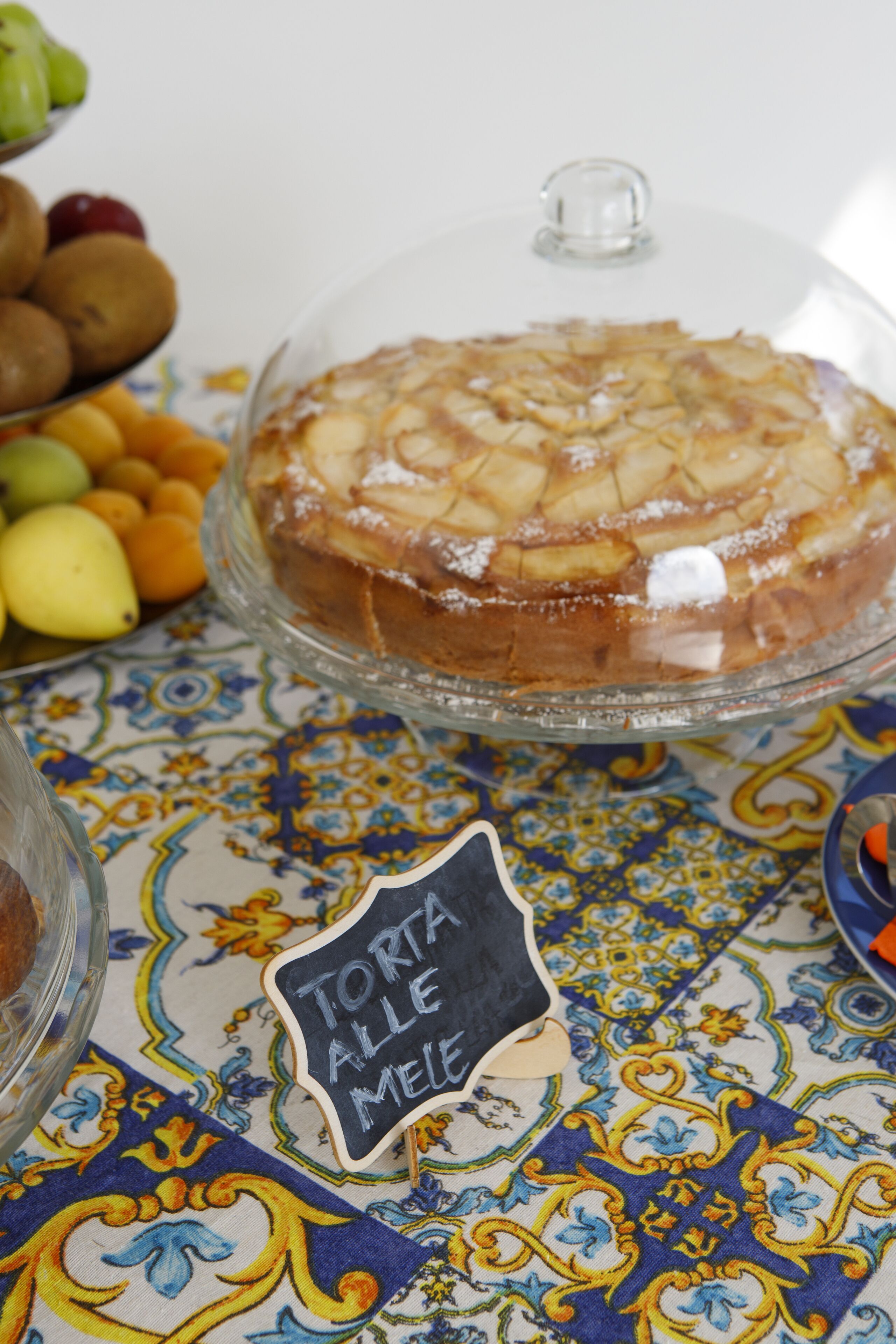 A dessert covered with a glass dome, a sign reading "TORTA ALLE MELE," and a tablecloth with a blue and yellow pattern.