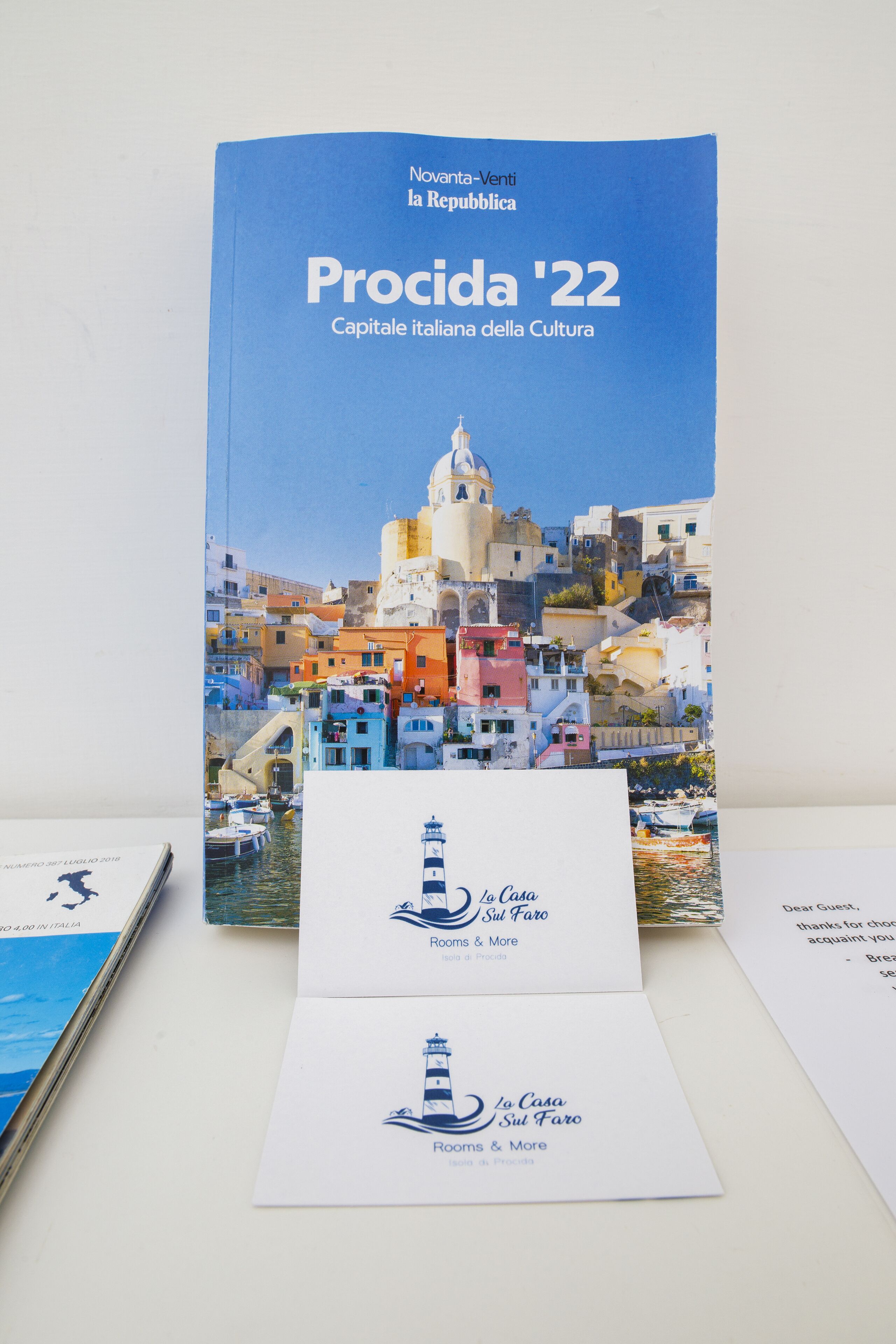 A brochure titled "Procida '22" with a scenic coastal town illustration. Below are two business cards for "La Casa Sul Faro" and "Rooms & More."