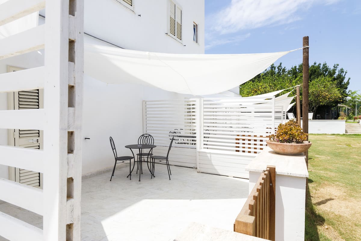 Un bâtiment blanc doté d’un patio couvert comprenant une table et des chaises, un escalier en bois et un pot de fleurs.