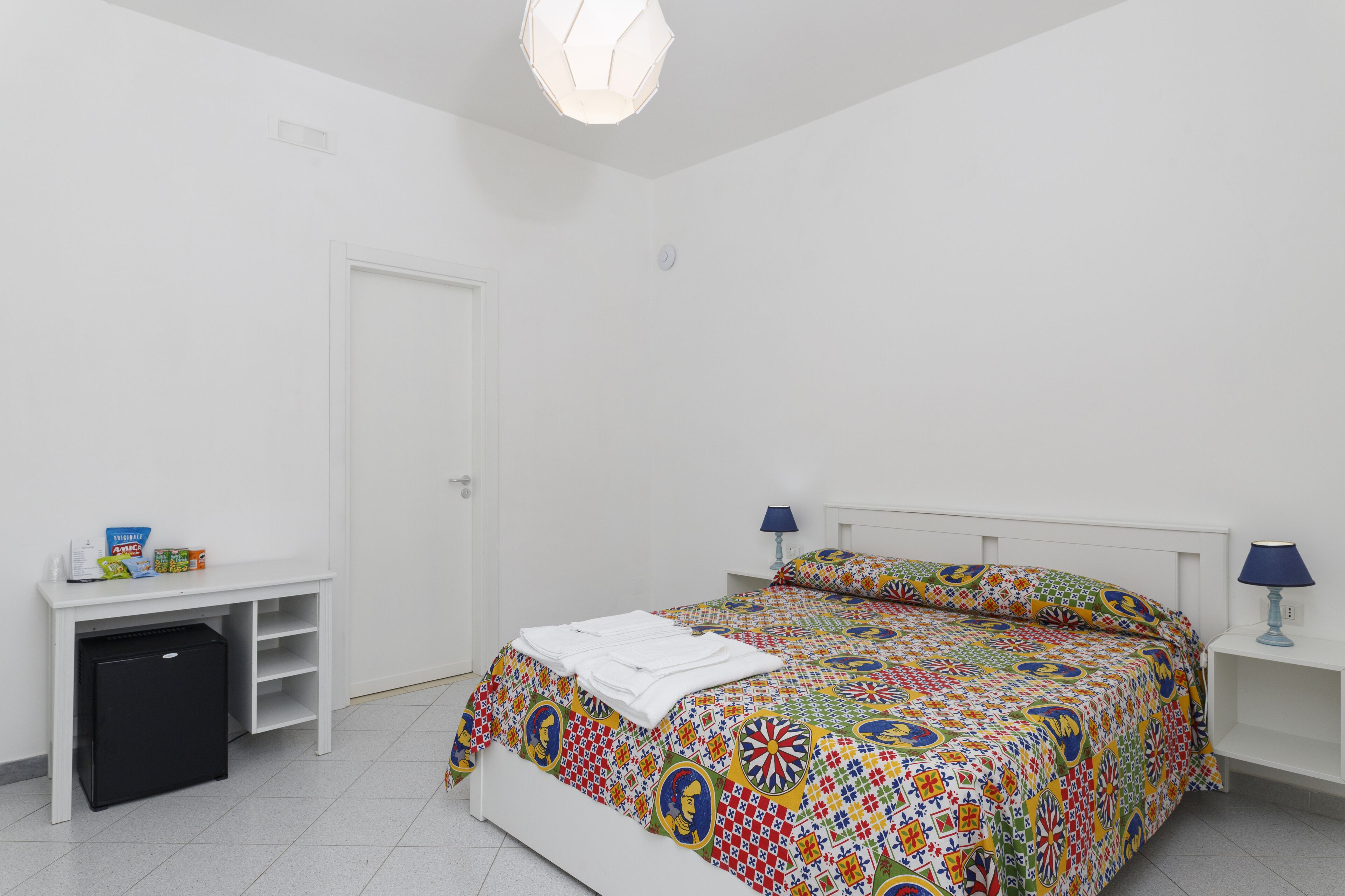 A bedroom with a bed, a desk, a small fridge, and a nightstand with a lamp.