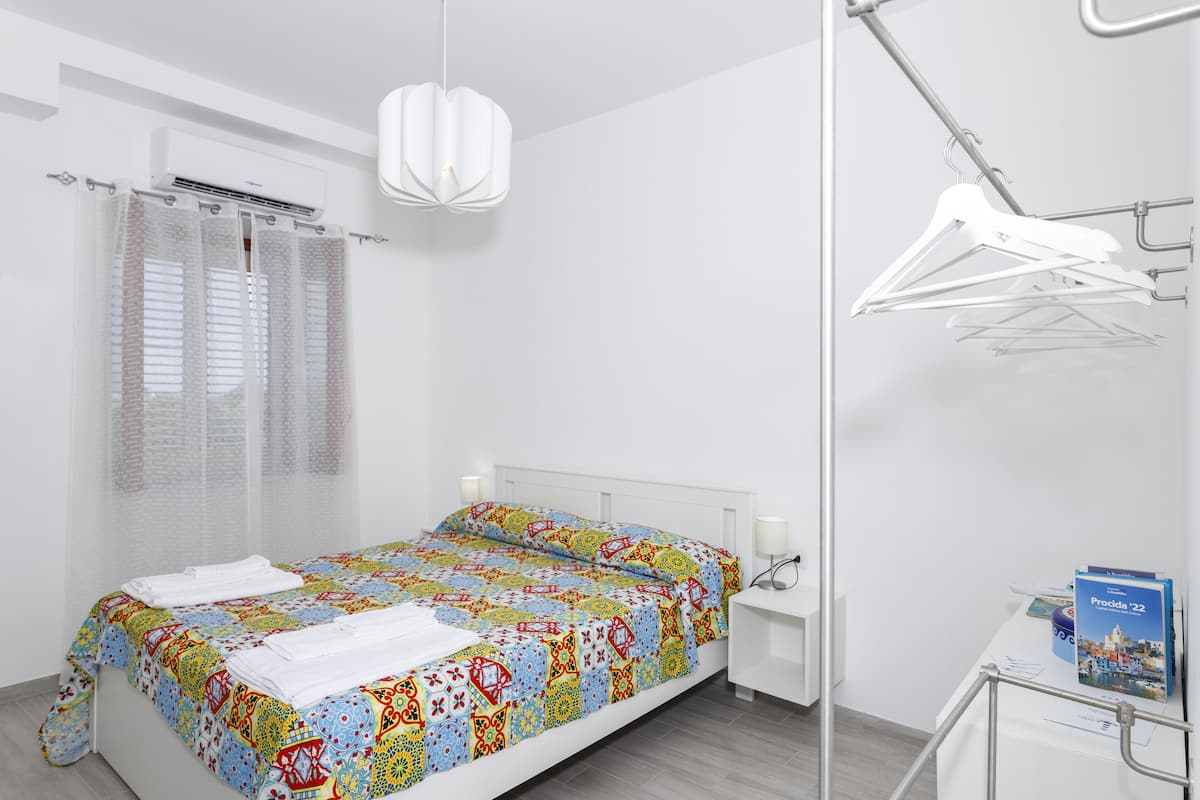 Une chambre d’hôtel moderne avec un lit soigneusement fait, une table de chevet avec une lampe, un luminaire suspendu, un porte-manteau et un petit bureau avec un livre et une carte.