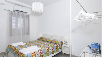 Una habitación de hotel moderna con una cama bien hecha, una mesita de noche con una lámpara, una lámpara colgante, un perchero y un escritorio pequeño con un libro y un mapa.