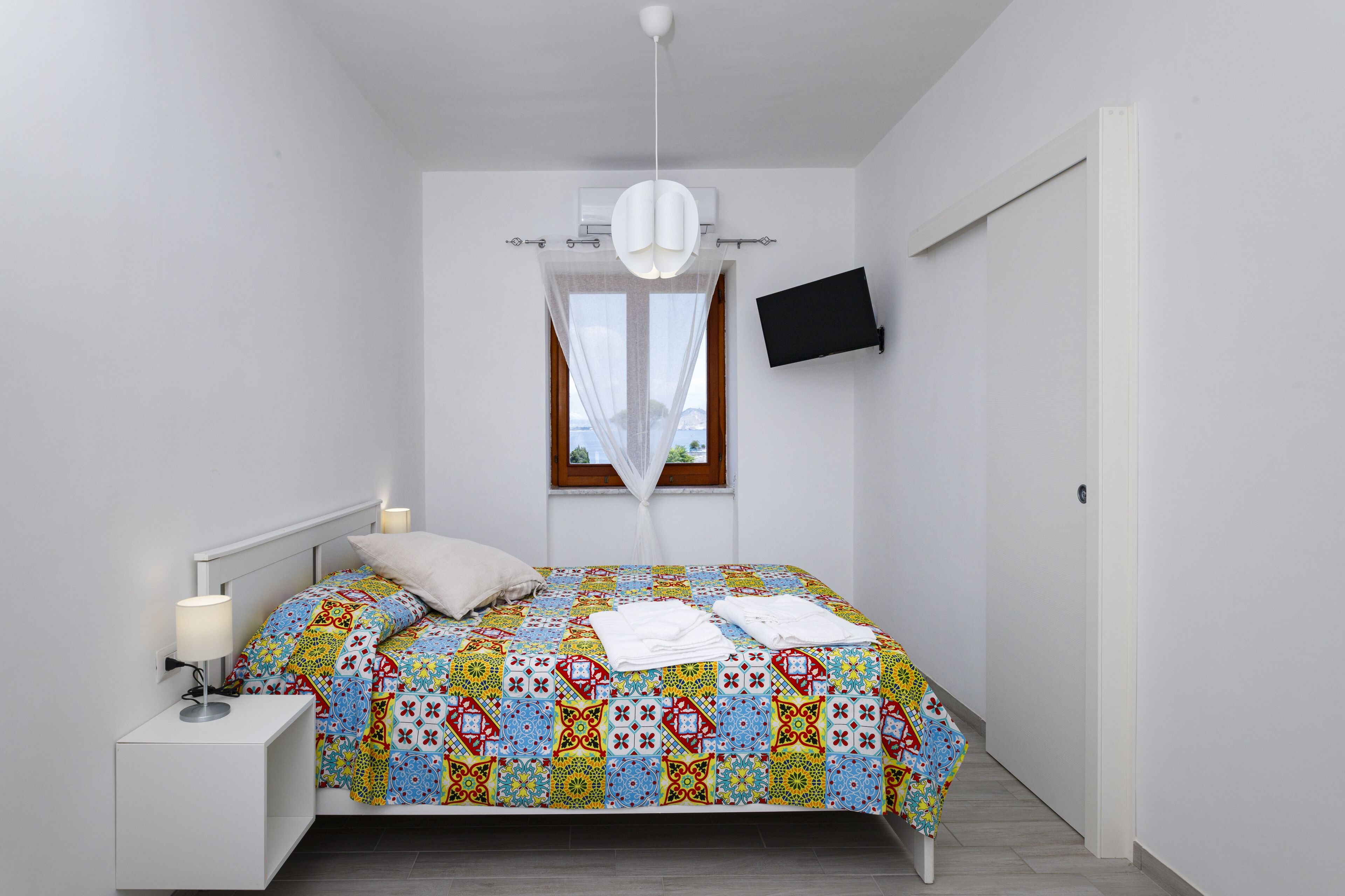 Une chambre à coucher comprenant un lit, une table de chevet, une lampe, un appareil de climatisation, une fenêtre et un téléviseur fixé au mur.
