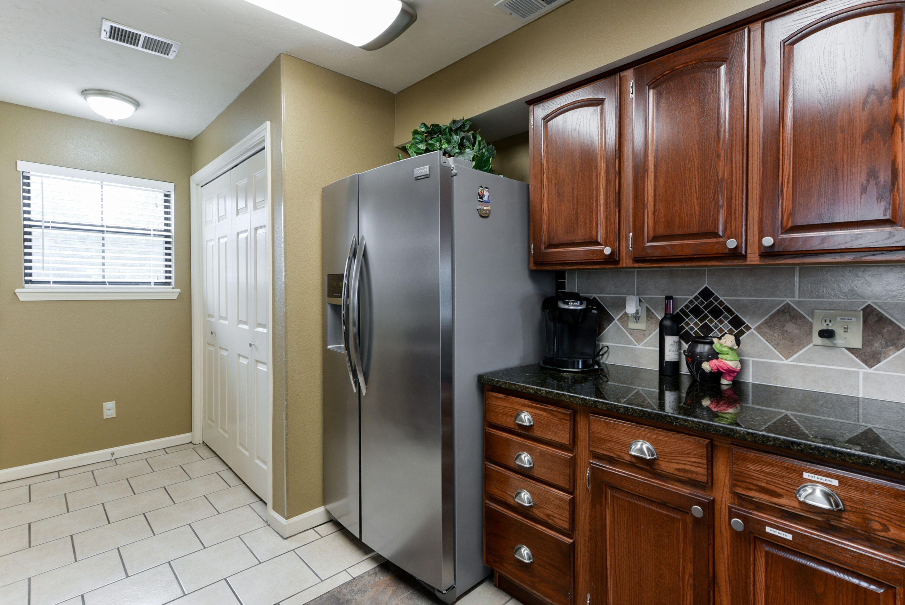A kitchen with wooden cabinets, a stainless steel refrigerator, a coffee maker, and a window with blinds.