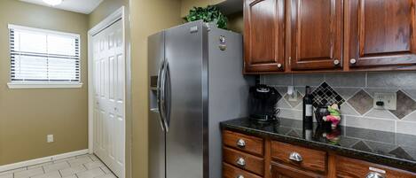 A kitchen with wooden cabinets, a stainless steel refrigerator, a coffee maker, and a window with blinds.