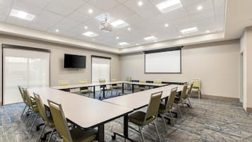Un salón de conferencias con una larga mesa, sillas, una pantalla de proyector y un televisor en la pared.