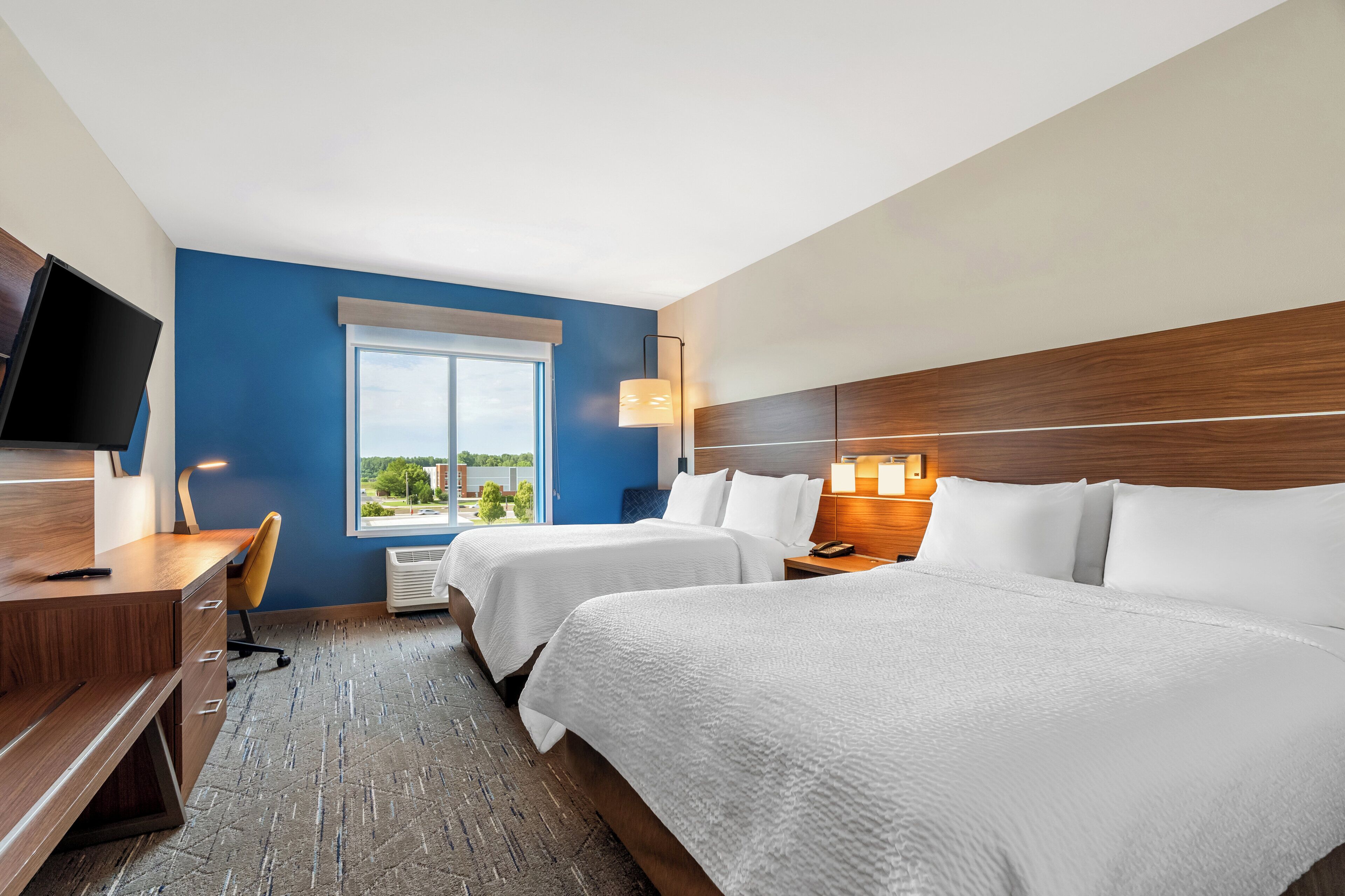 A hotel room with two beds, a desk, a television, and a window with a view of buildings and trees.