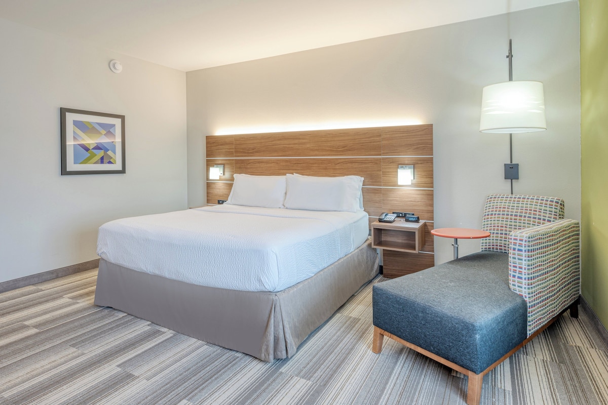 Ein Hotelzimmer mit Bett, Nachttischen, einer Lampe, einem Sessel und einem gerahmten Bild an der Wand.