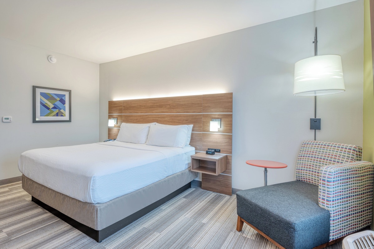 Ein modernes Hotelzimmer mit einem großen Bett, einem Nachttisch, einem kleinen Tisch, einem gemusterten Sessel und einer Wandlampe.
