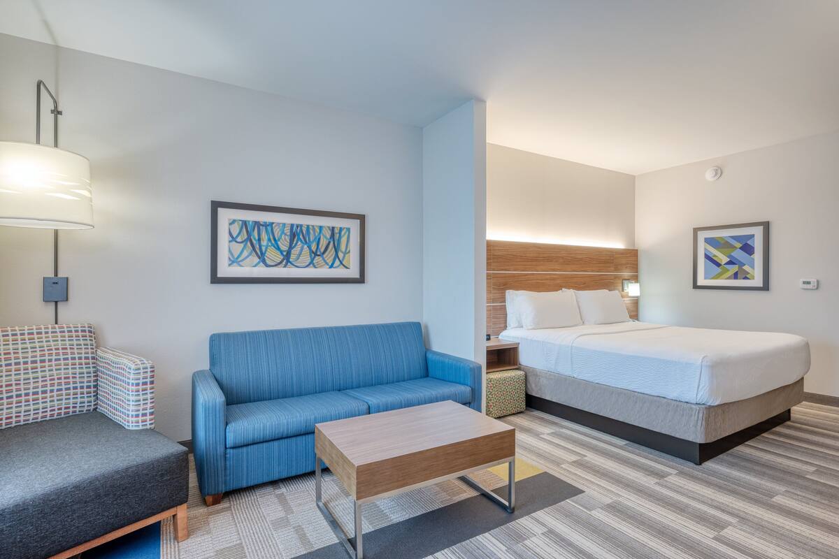 A hotel room with a blue sofa, a bed, a wooden coffee table, and two abstract paintings on the walls.