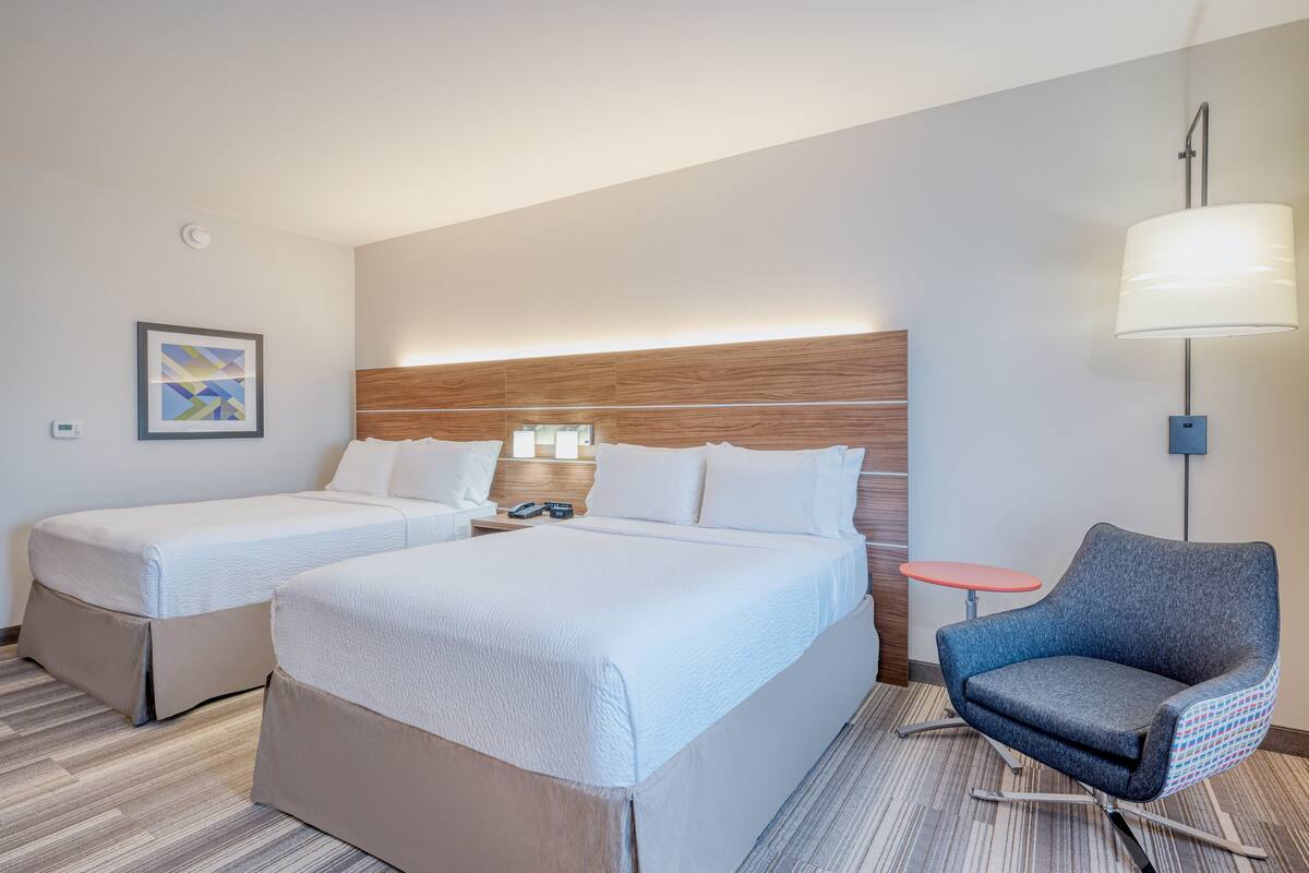 A hotel room with two beds, a nightstand, a lamp, a chair, and a small table.