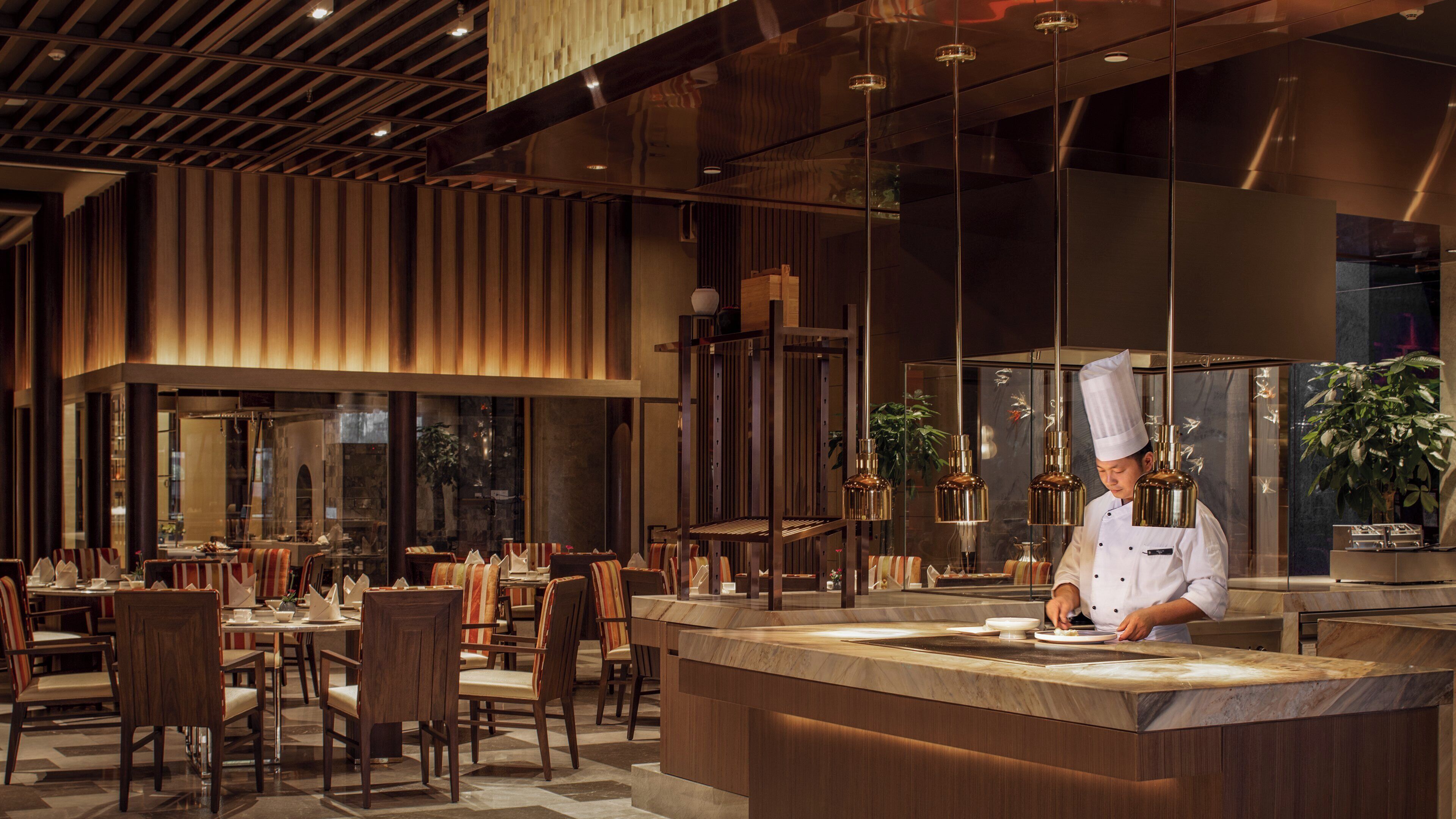 A modern restaurant interior with a chef preparing food at a counter, wooden paneling on the walls, and a large window overlooking the dining area.