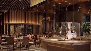A modern restaurant interior with a chef preparing food at a counter, wooden paneling on the walls, and a large window overlooking the dining area. - InterContinental Tianjin Yujiapu Hotel & Residences by IHG (Tianjin)